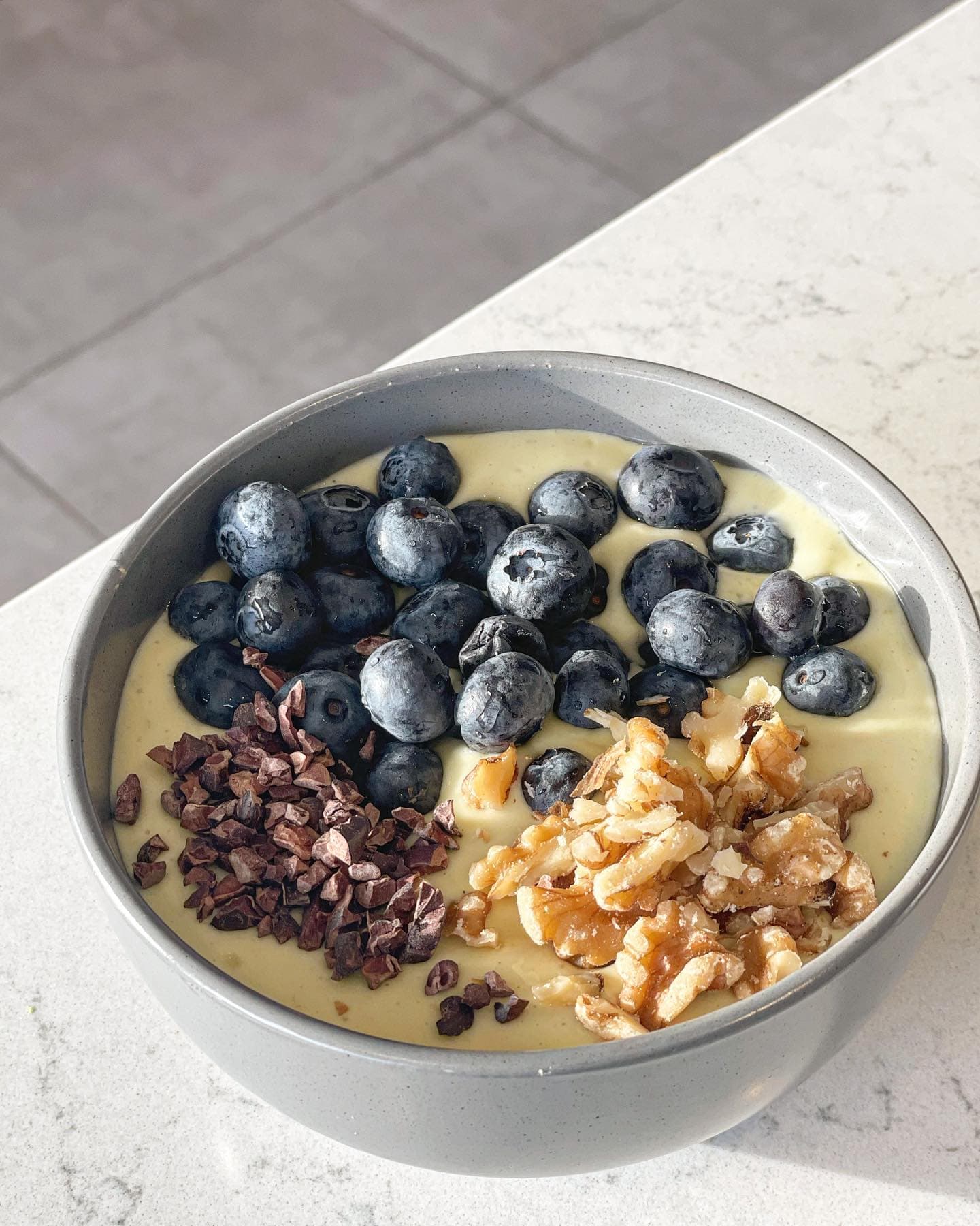 En grå skål med ljusgul smoothie på en marmorbänk, toppad med blåbär, hackade valnötter och kakaonibs.