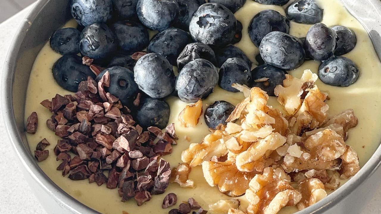 En grå skål med ljusgul smoothie på en marmorbänk, toppad med blåbär, hackade valnötter och kakaonibs.