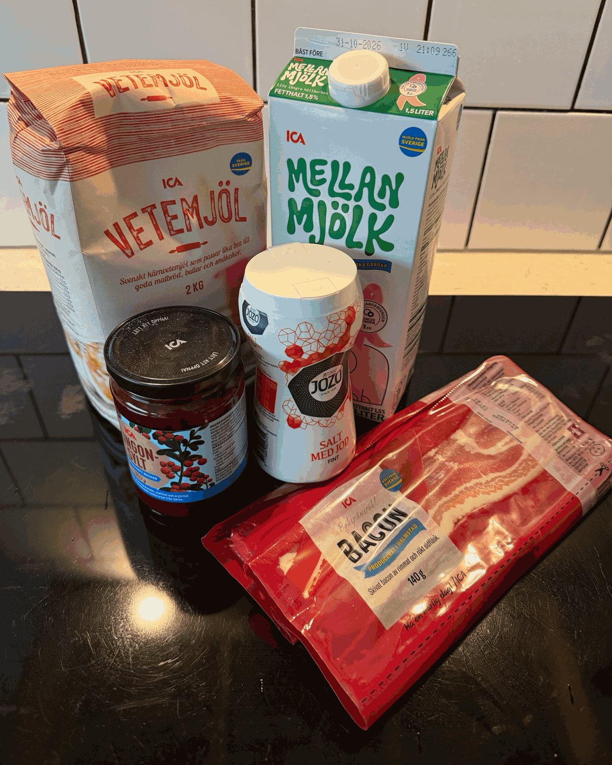 Vetemjöl, mjölk, lingonsylt, salt och ett paket bacon står uppställda på en mörk bänkskiva.