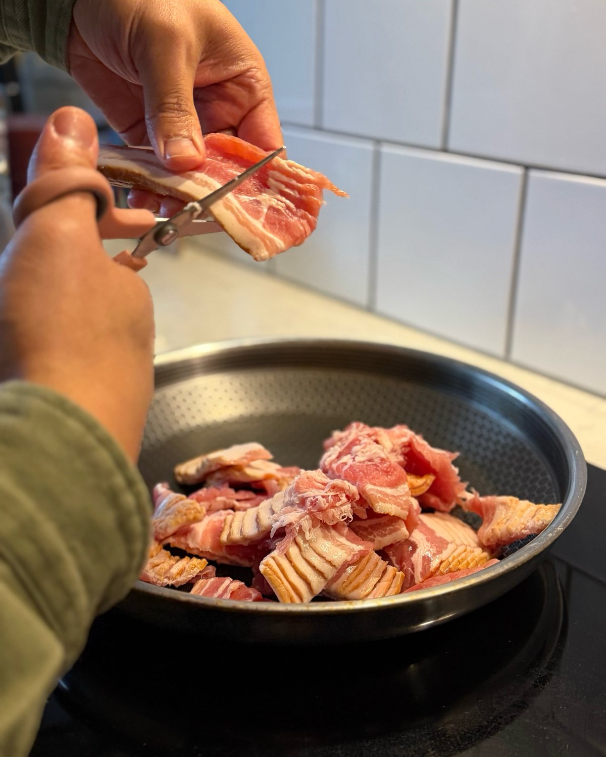 Två händer klipper en baconskiva med en sax rakt ner i en stekpanna som redan är halvfull.