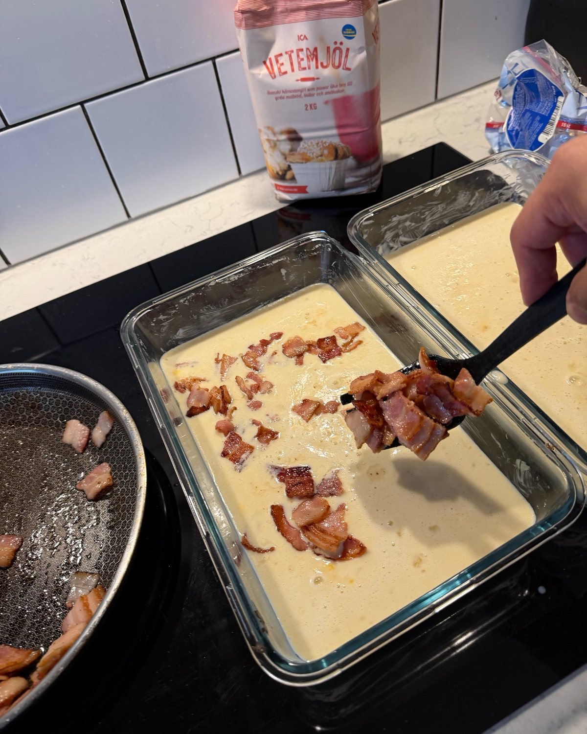 En stekspade lyfter stekta baconbitar från pannan ner i en glasform med ljus smet.