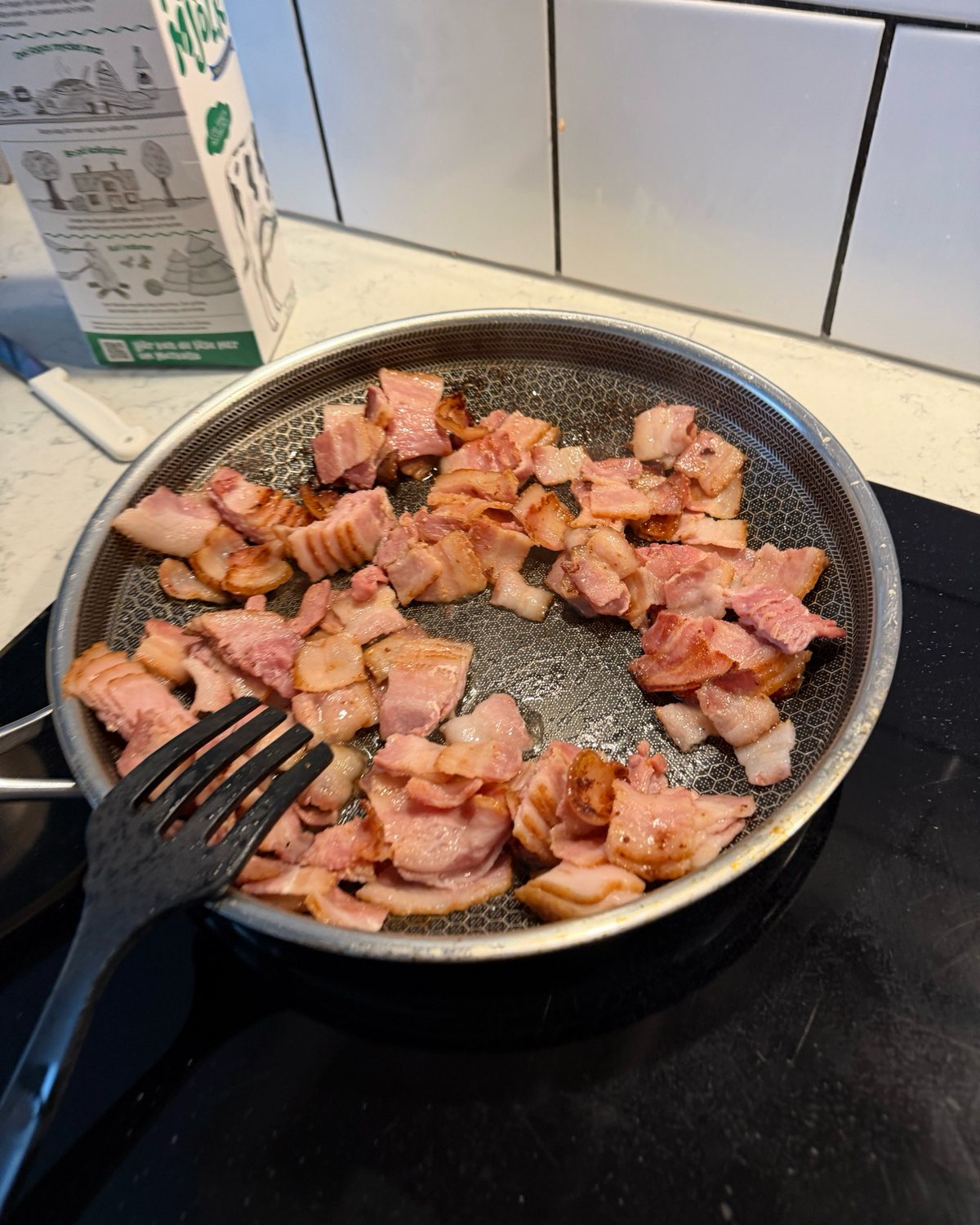 Baconbitar i en stekpanna, några gyllenbruna och några fortfarande ljusa, med en stekspade i kanten.